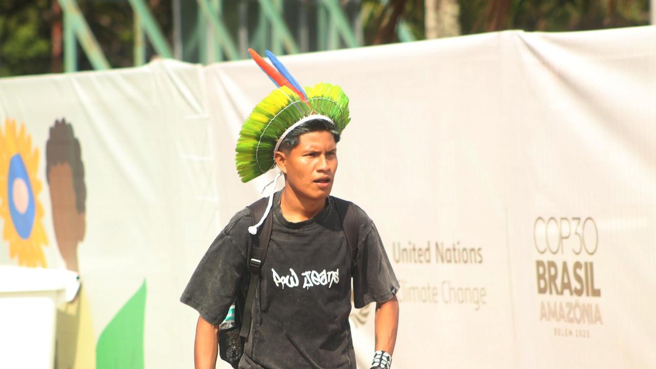 Eine indigene Person geht mit einem traditionellen Kopfschmuck an dem Logo der COP30 in Belém, Brasilien, vorbei