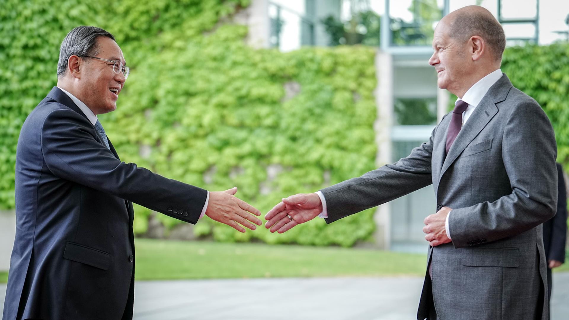 Bundeskanzler Scholz empfängt Chinas Ministerpräsident Li Qiang vor dem Bundeskanzleramt. Beide strecken die Hand aus, um sich die Hände zu schütteln und lächeln einander an.