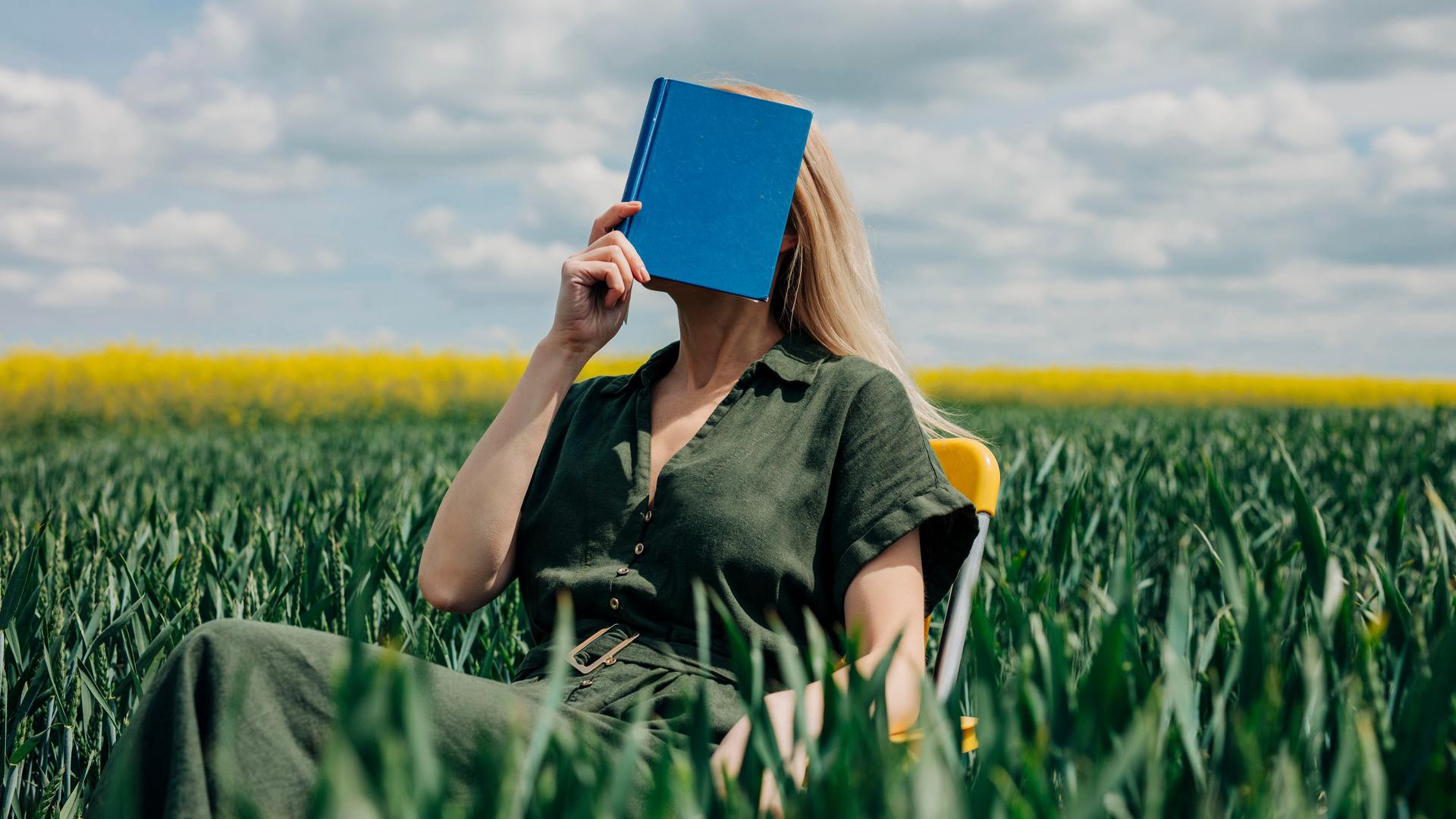 Eine Frau sitzt mit einem blauen Buch vor dem Gesicht in einem Feld auf einem Stuhl. Eine Frau sitzt mit einem blauen Buch vor dem Gesicht in einem Feld auf einem Stuhl.