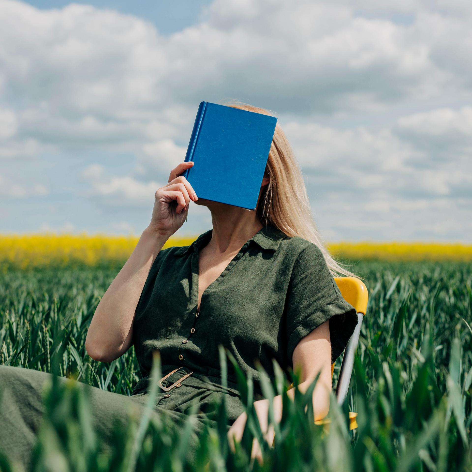 Eine Frau sitzt mit einem blauen Buch vor dem Gesicht in einem Feld auf einem Stuhl.
