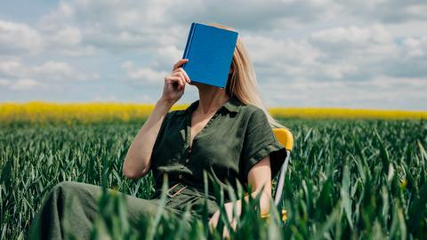 Eine Frau sitzt mit einem blauen Buch vor dem Gesicht in einem Feld auf einem Stuhl.