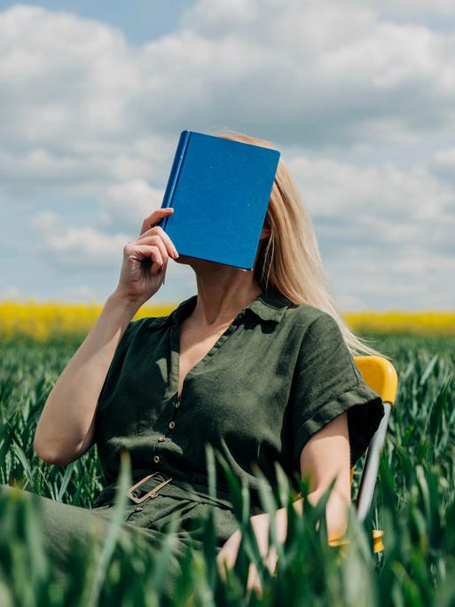Eine Frau sitzt mit einem blauen Buch vor dem Gesicht in einem Feld auf einem Stuhl.