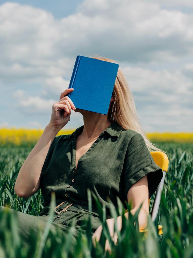 Eine Frau sitzt mit einem blauen Buch vor dem Gesicht in einem Feld auf einem Stuhl.