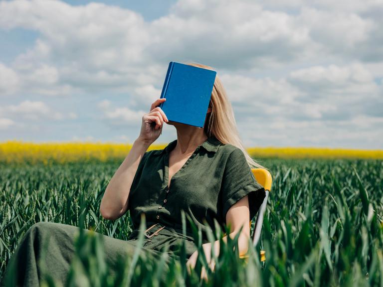 Eine Frau sitzt mit einem blauen Buch vor dem Gesicht in einem Feld auf einem Stuhl.
