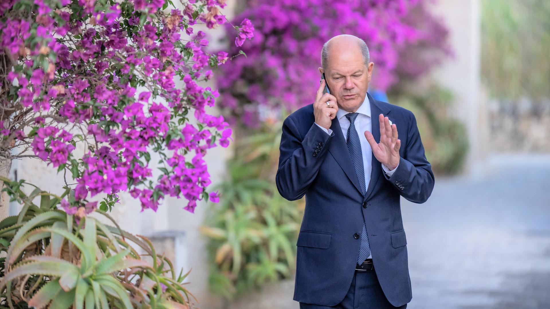 Scholz läuft an einer mit vielen lila Blumen behangenen Wand entlang. Er hält das Handy ans Ohr und gestikuliert mit der linken Hand.