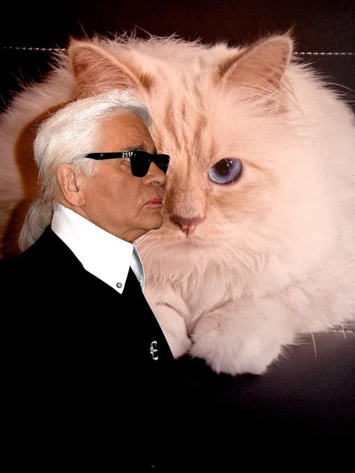 Karl Lagerfeld mit Sonnenbrille und Pferdeschwanz steht bei einer Vernissage vor einem großen Bild seiner Katze Choupette