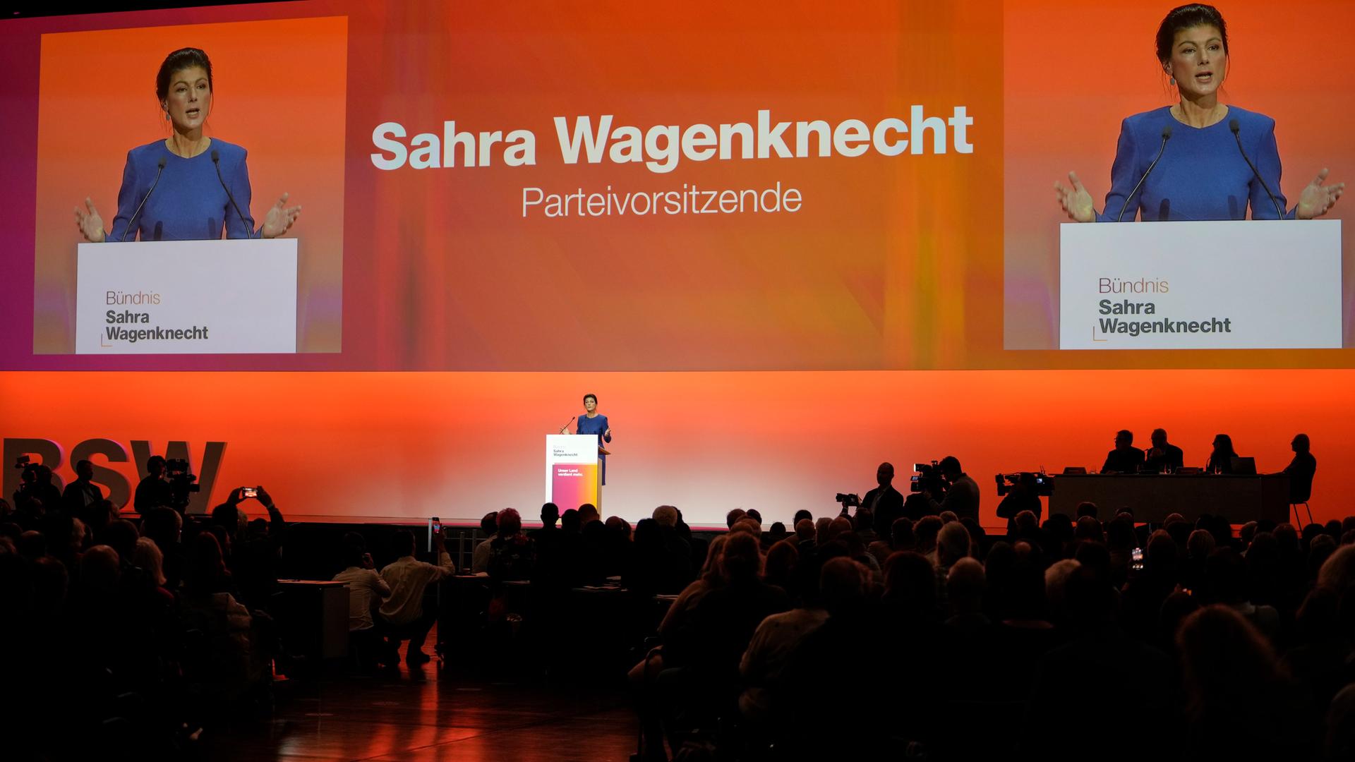 Sahra Wagenknecht steht in einem blauen Kleid beim Parteitag auf der Bühne und spricht vor einer orangefarbenen Wand in ein Mikrofon; vor der Bühne sind zahlreiche Delegierte zu sehen. 