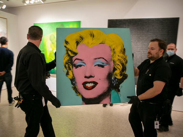 Das Bild "Shot Sage Blue Marilyn" wird von zwei Arbeitern durch eine Galerie getragen.