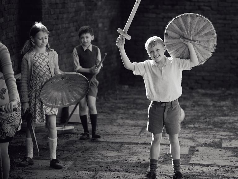 Im Still aus "Belfast" steht der kindliche Hauptdarsteller mit einem aus Holz gebastelten Schild und Schwert jubelnd auf einem Hof.
