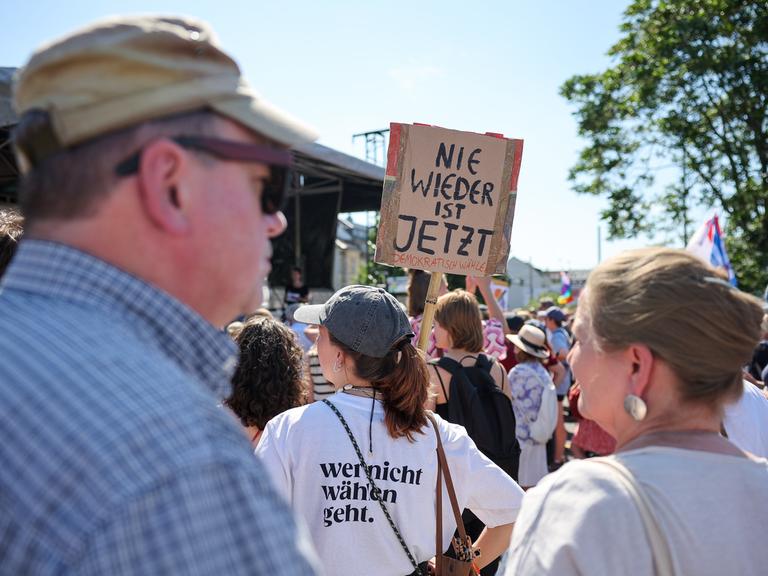 Eine Teilnehmerin hält ein Schild "Nie wieder ist jetzt" auf einer Kundgebung in die Höhe. Andere Menschen sind ebenfalls im Bild