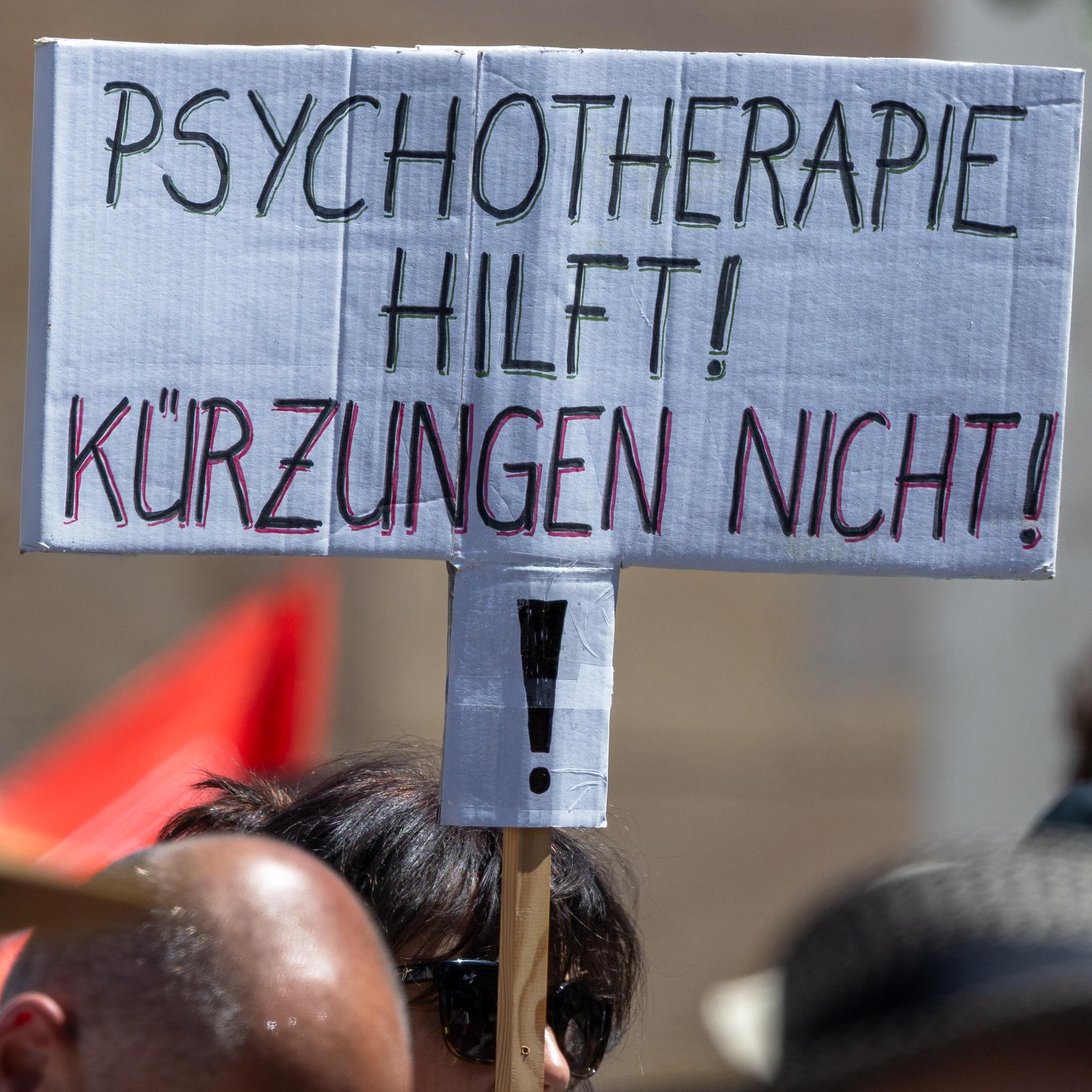 Über den Köpfen mehrerer Demonstrierender ist ein handgeschriebenes Schild zu sehen. Darauf steht in schwarzen und roten Großbuchstaben: „Psychotherapie hilft! Kürzungen nicht!“ Die Menschen im Vordergrund sind unscharf abgebildet.