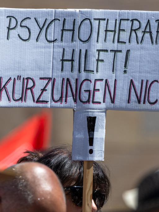 Über den Köpfen mehrerer Demonstrierender ist ein handgeschriebenes Schild zu sehen. Darauf steht in schwarzen und roten Großbuchstaben: „Psychotherapie hilft! Kürzungen nicht!“ Die Menschen im Vordergrund sind unscharf abgebildet. Über den Köpfen mehrerer Demonstrierender ist ein handgeschriebenes Schild zu sehen. Darauf steht in schwarzen und roten Großbuchstaben: „Psychotherapie hilft! Kürzungen nicht!“ Die Menschen im Vordergrund sind unscharf abgebildet.