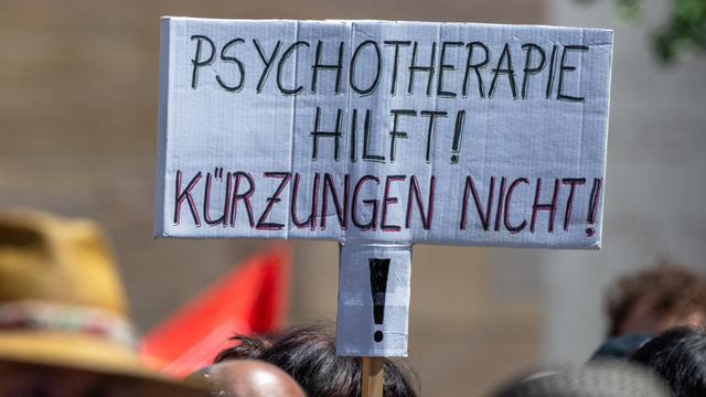 Über den Köpfen mehrerer Demonstrierender ist ein handgeschriebenes Schild zu sehen. Darauf steht in schwarzen und roten Großbuchstaben: „Psychotherapie hilft! Kürzungen nicht!“ Die Menschen im Vordergrund sind unscharf abgebildet.