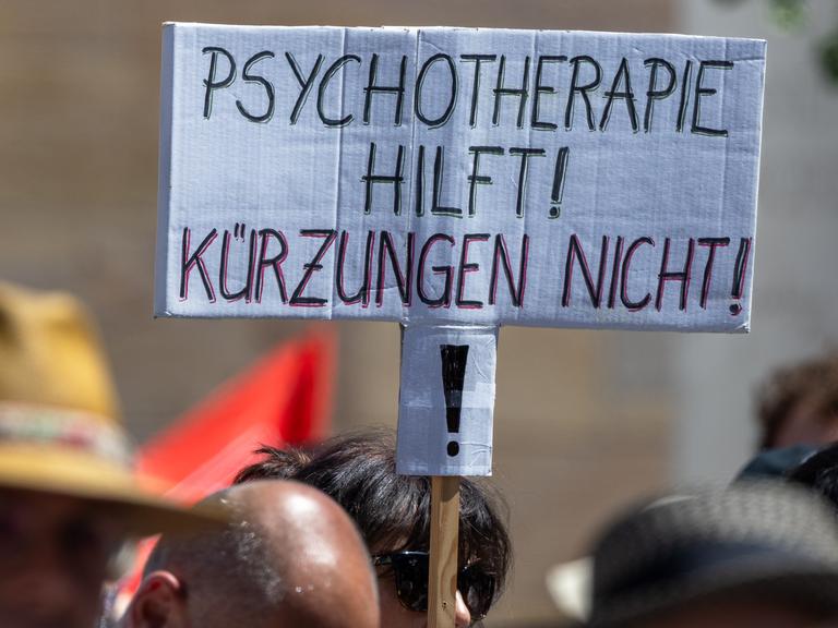 Über den Köpfen mehrerer Demonstrierender ist ein handgeschriebenes Schild zu sehen. Darauf steht in schwarzen und roten Großbuchstaben: „Psychotherapie hilft! Kürzungen nicht!“ Die Menschen im Vordergrund sind unscharf abgebildet. Über den Köpfen mehrerer Demonstrierender ist ein handgeschriebenes Schild zu sehen. Darauf steht in schwarzen und roten Großbuchstaben: „Psychotherapie hilft! Kürzungen nicht!“ Die Menschen im Vordergrund sind unscharf abgebildet.