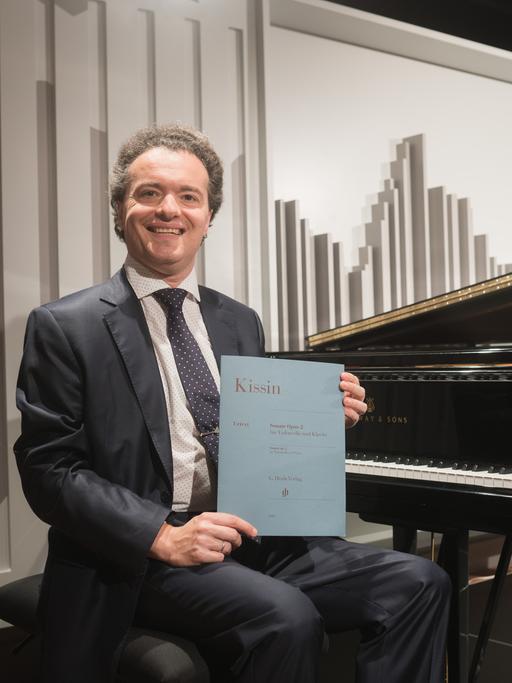Der Pianist Evgeny Kissin sitzt auf einem Klavierhocker, hinter ihm steht ein großer Flügel. Er zeigt stolz eine Notenausgabe, es ist ein von ihm komponiertes Werk.