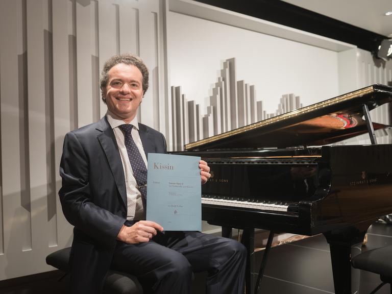 Der Pianist Evgeny Kissin sitzt auf einem Klavierhocker, hinter ihm steht ein großer Flügel. Er zeigt stolz eine Notenausgabe, es ist ein von ihm komponiertes Werk.