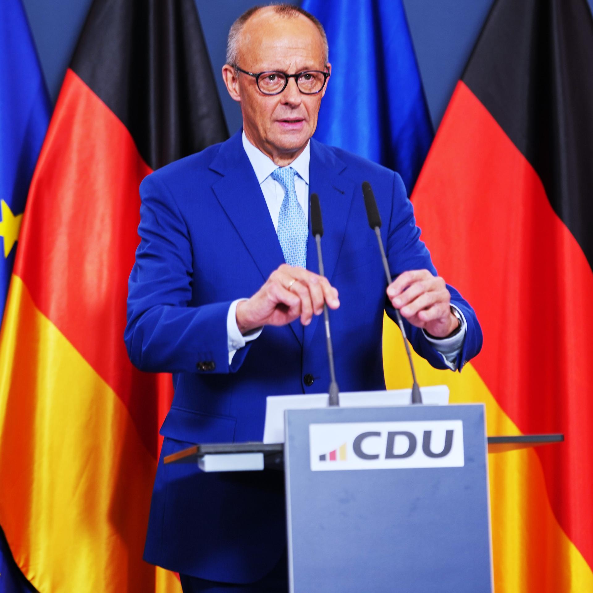 Bundeskanzler Friedrich Merz steht an einem Rednerpult und rückt zwei Mikrofone zurecht. Im Hintergrund sind die Flaggen der Bundesrepublik und der Europäischen Union aufgestellt. 