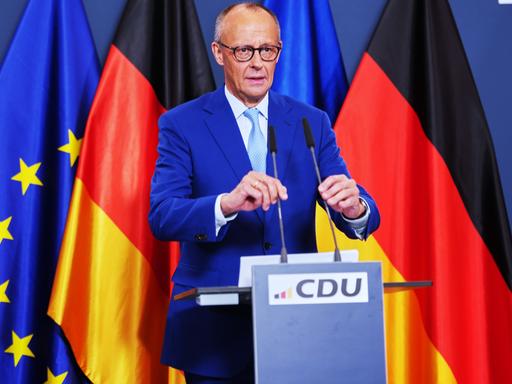 Bundeskanzler Friedrich Merz steht an einem Rednerpult und rückt zwei Mikrofone zurecht. Im Hintergrund sind die Flaggen der Bundesrepublik und der Europäischen Union aufgestellt. 