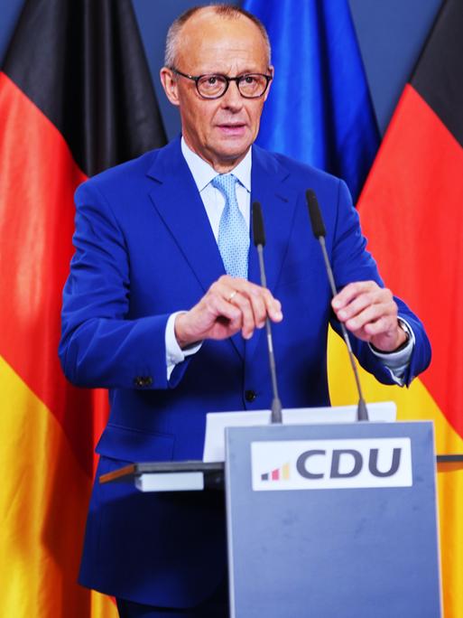 Bundeskanzler Friedrich Merz steht an einem Rednerpult und rückt zwei Mikrofone zurecht. Im Hintergrund sind die Flaggen der Bundesrepublik und der Europäischen Union aufgestellt. 