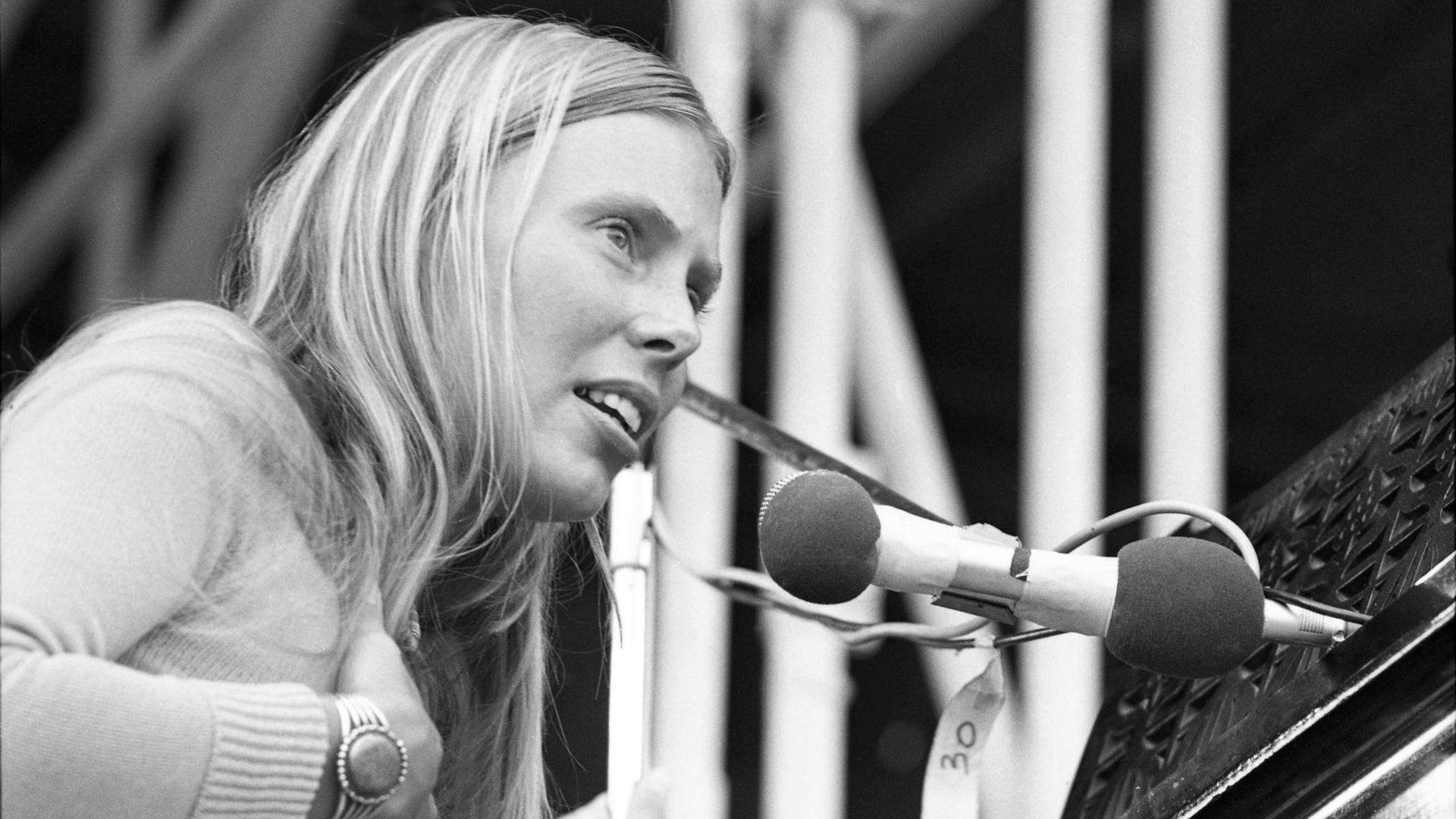 Die Schwarzweiß-Fotografie zeigt Joni Mitchell im Jahr 1970 beim Isle of Wight Festival - sie trägt lange, blonde Haare, fasst sich mit der rechten Hand auf die Brust und singt in ein Mikrofon.