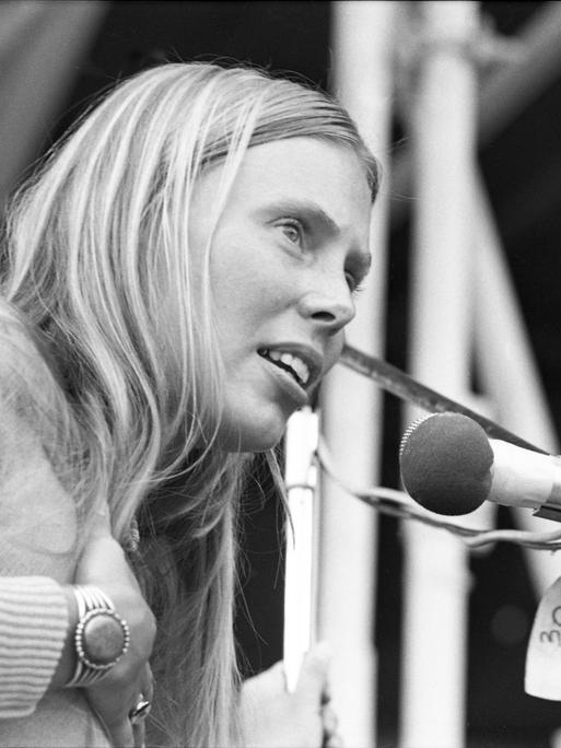 Die Schwarzweiß-Fotografie zeigt Joni Mitchell im Jahr 1970 beim Isle of Wight Festival - sie trägt lange, blonde Haare, fasst sich mit der rechten Hand auf die Brust und singt in ein Mikrofon.