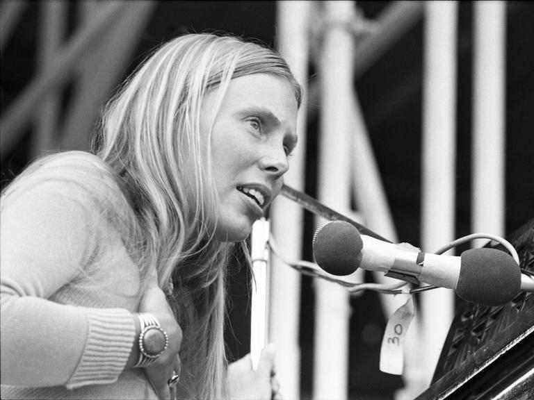 Die Schwarzweiß-Fotografie zeigt Joni Mitchell im Jahr 1970 beim Isle of Wight Festival - sie trägt lange, blonde Haare, fasst sich mit der rechten Hand auf die Brust und singt in ein Mikrofon.