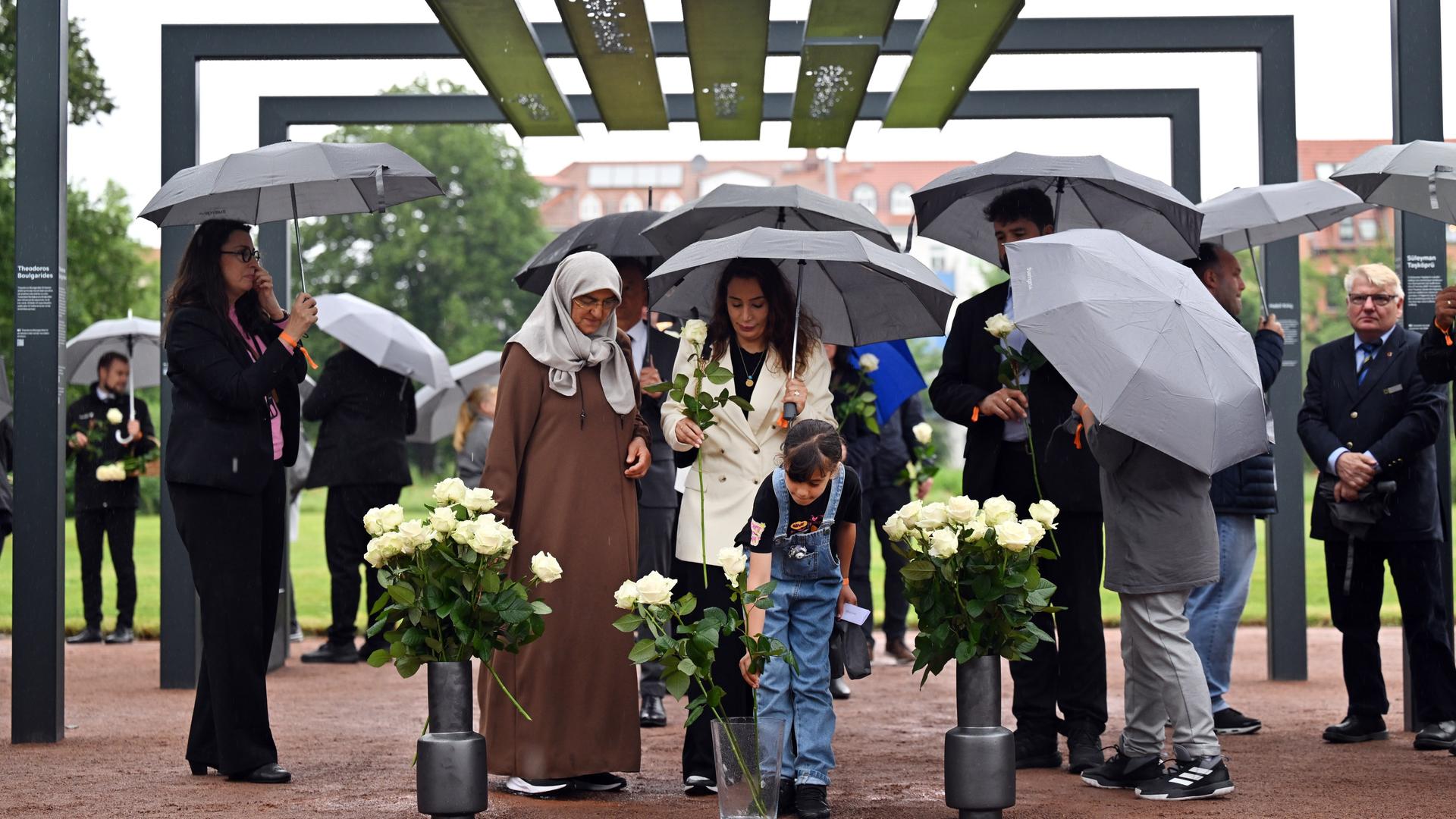 Mehrere Menschen mit Schirmen stehen unter einem aus Stahlbögen bestehenden Mahnmal, an denen die Namen und Biografien der NSU-Opfer zu lesen sind. Mehrere Menschen mit Schirmen stehen unter einem aus Stahlbögen bestehenden Mahnmal, an denen die Namen und Biografien der NSU-Opfer zu lesen sind.