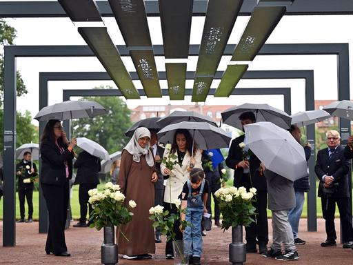 Mehrere Menschen mit Schirmen stehen unter einem aus Stahlbögen bestehenden Mahnmal, an denen die Namen und Biografien der NSU-Opfer zu lesen sind. Mehrere Menschen mit Schirmen stehen unter einem aus Stahlbögen bestehenden Mahnmal, an denen die Namen und Biografien der NSU-Opfer zu lesen sind.