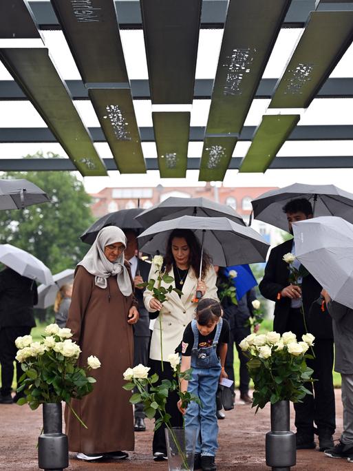 Mehrere Menschen mit Schirmen stehen unter einem aus Stahlbögen bestehenden Mahnmal, an denen die Namen und Biografien der NSU-Opfer zu lesen sind.
