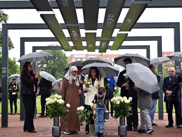 Mehrere Menschen mit Schirmen stehen unter einem aus Stahlbögen bestehenden Mahnmal, an denen die Namen und Biografien der NSU-Opfer zu lesen sind.