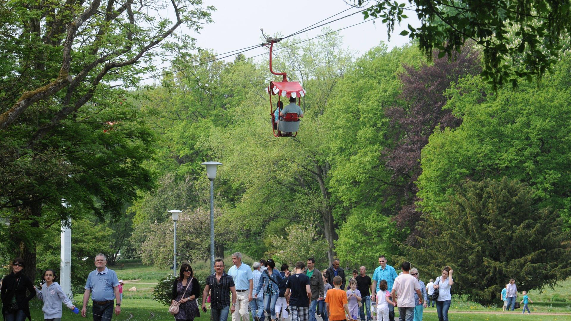 Eine Gondel der Seilbahn im Deutsch-Französischen-Garten in Saarbrücken schwebt über einem Weg, auf dem viele Menschen spazierengehen.