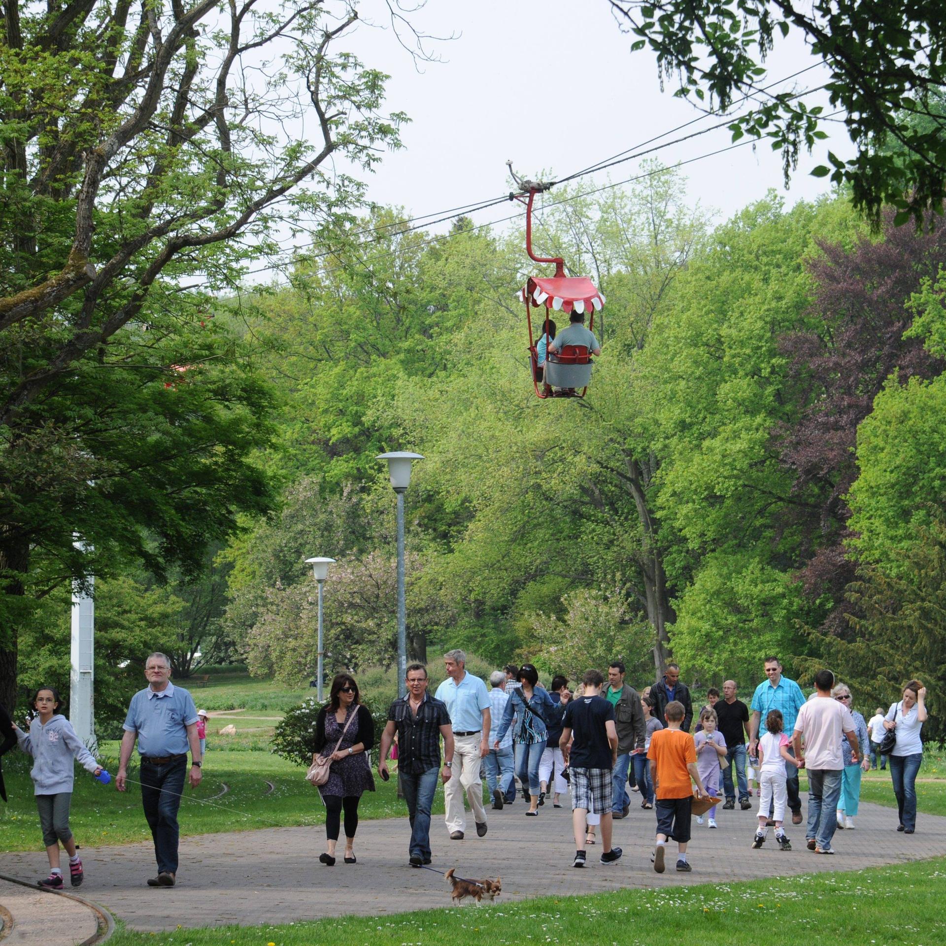 Eine Gondel der Seilbahn im Deutsch-Französischen-Garten in Saarbrücken schwebt über einem Weg, auf dem viele Menschen spazierengehen.