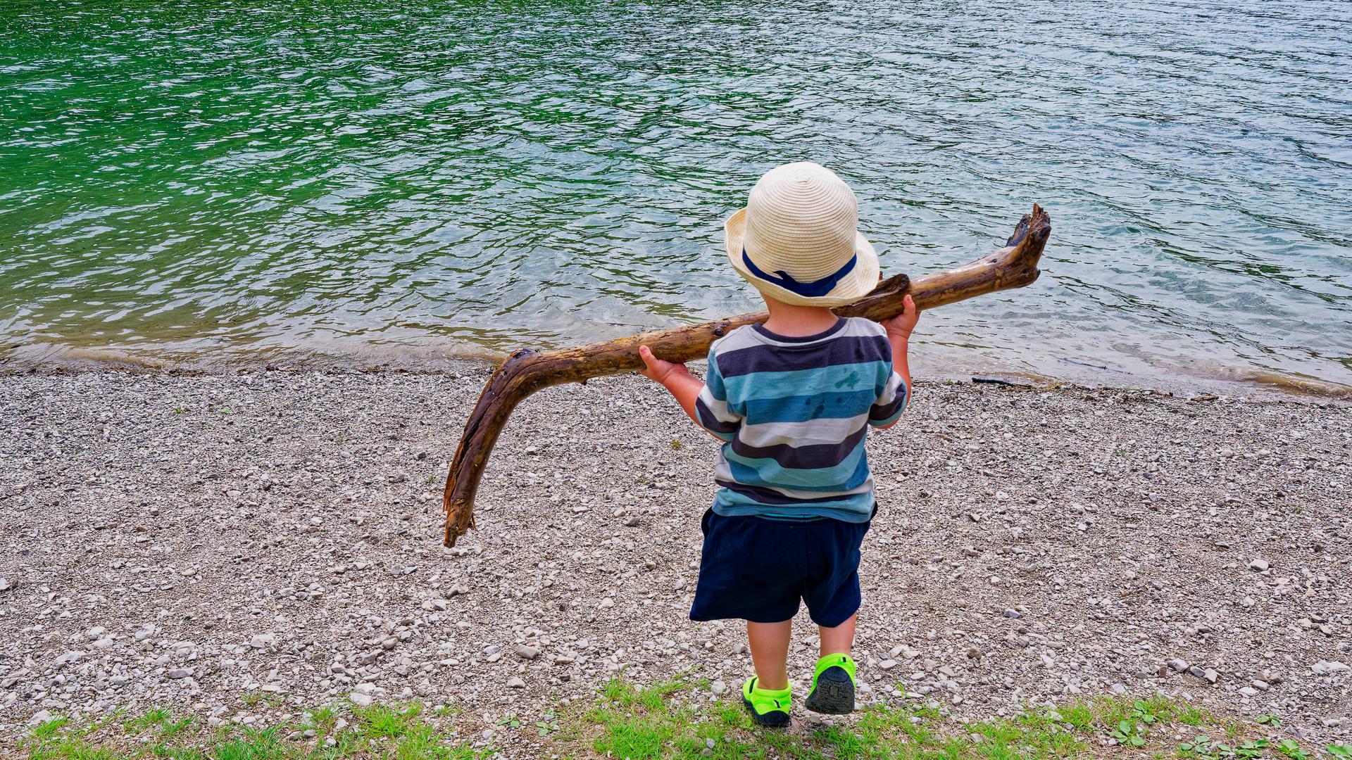 Ein kleines Kind steht mit dem Rücken zum Betrachter am Ufer eines Gewässers und hält mit beiden Händen einen großen Stock hoch