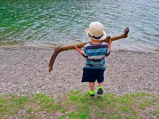 Ein kleines Kind steht mit dem Rücken zum Betrachter am Ufer eines Gewässers und hält mit beiden Händen einen großen Stock hoch