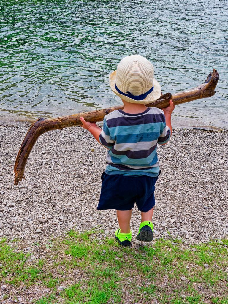 Ein kleines Kind steht mit dem Rücken zum Betrachter am Ufer eines Gewässers und hält mit beiden Händen einen großen Stock hoch