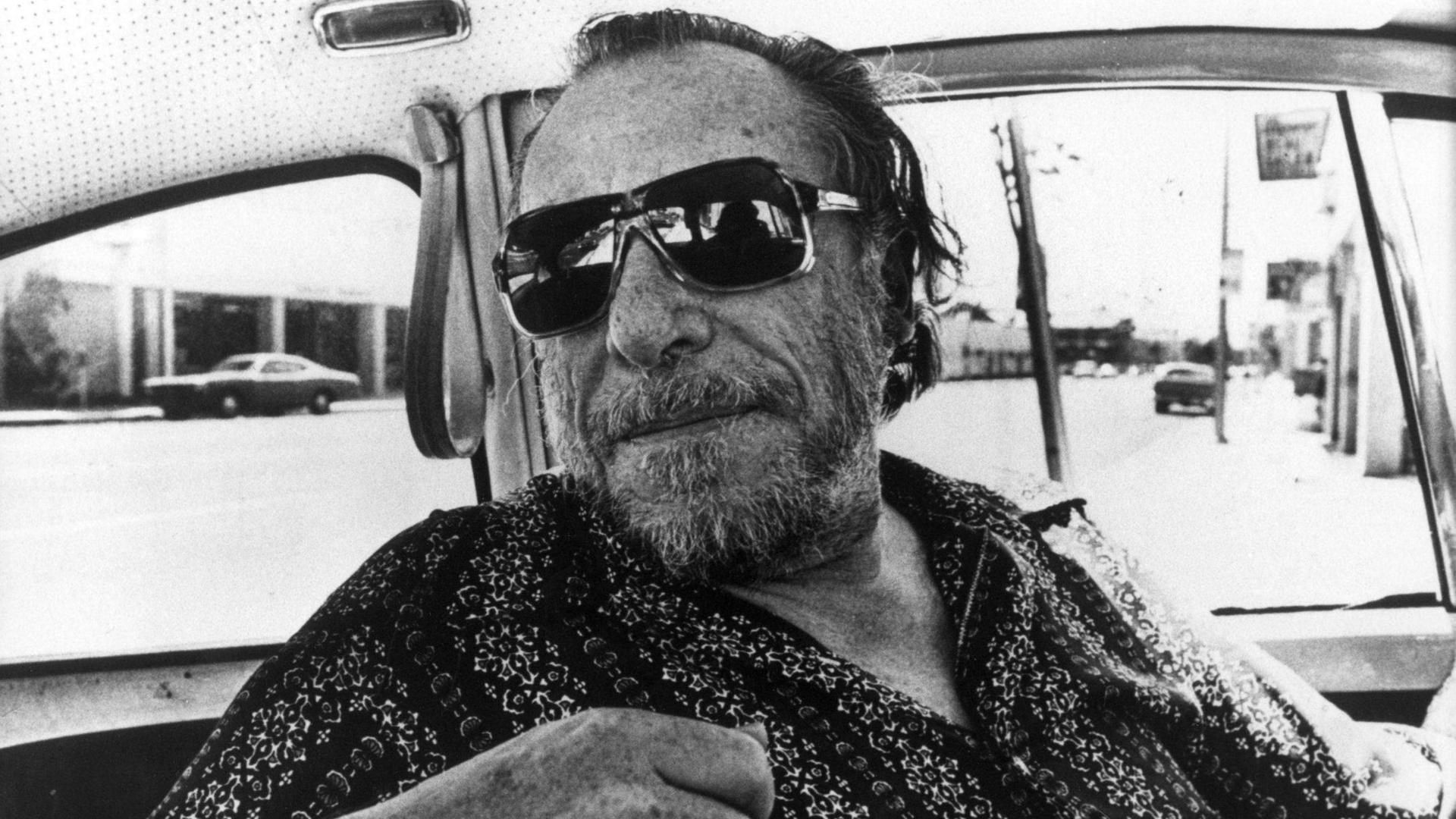Charles Bukowski trägt eine coole Sonnenbrille und blickt lässig über einen Autositz ins Hintere der Autokabine Charles Bukowski trägt eine coole Sonnenbrille und blickt lässig über einen Autositz ins Hintere der Autokabine