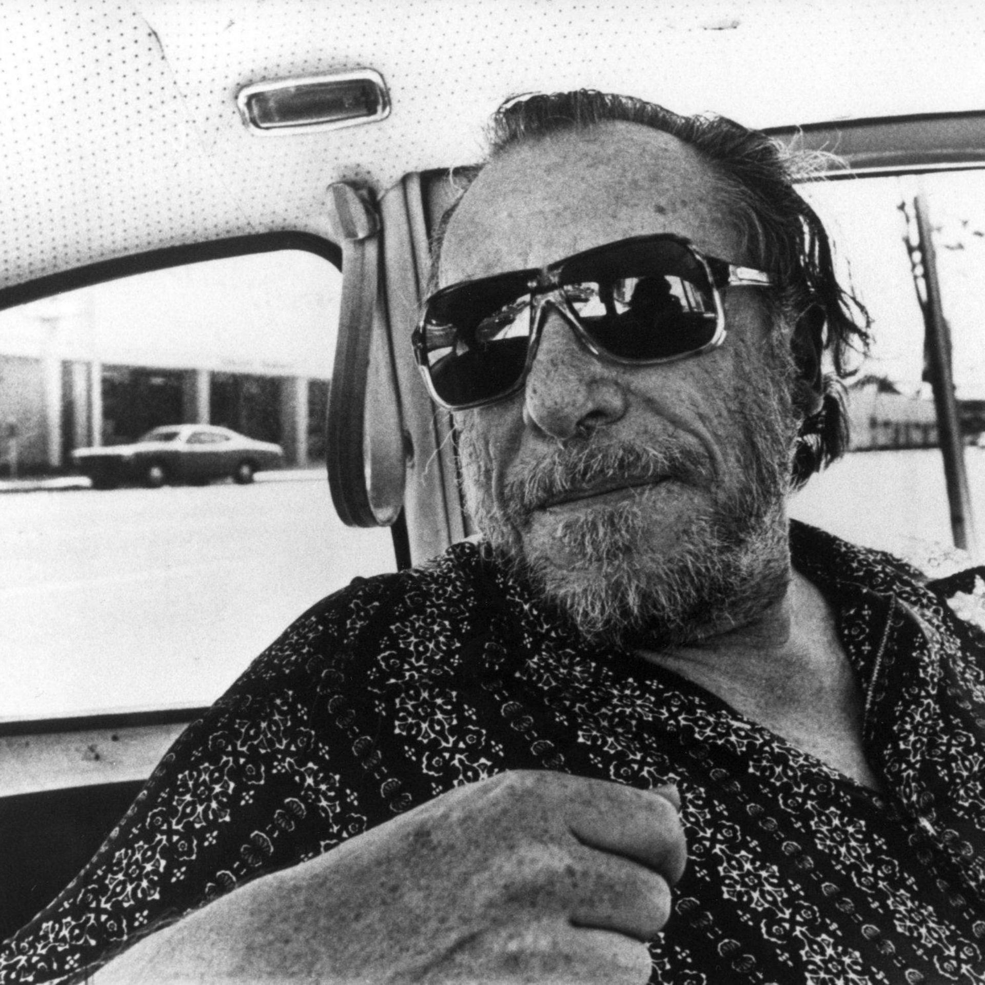 Charles Bukowski trägt eine coole Sonnenbrille und blickt lässig über einen Autositz ins Hintere der Autokabine