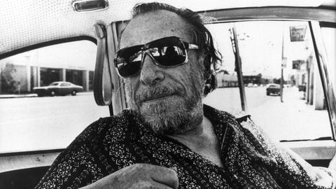 Charles Bukowski trägt eine coole Sonnenbrille und blickt lässig über einen Autositz ins Hintere der Autokabine
