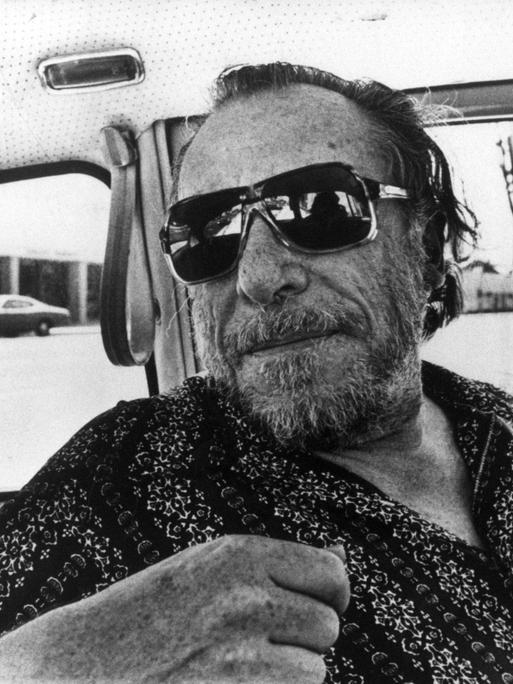 Charles Bukowski trägt eine coole Sonnenbrille und blickt lässig über einen Autositz ins Hintere der Autokabine