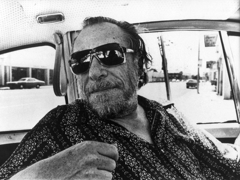 Charles Bukowski trägt eine coole Sonnenbrille und blickt lässig über einen Autositz ins Hintere der Autokabine