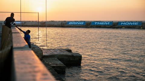Zwei Fischer und ein Taucher im Hafen von Jaffa am 1. November 2023. Auf der gegenüberliegenden Seite lässt sich auf einer Mauer in großen Lettern "Bring them home" - "Bringt sie heim" lesen.
