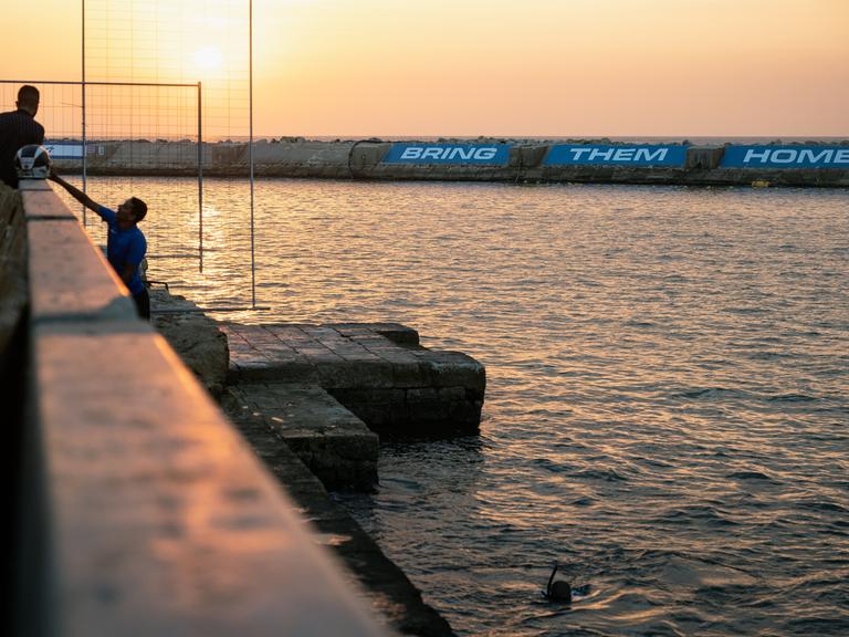 Zwei Fischer und ein Taucher im Hafen von Jaffa am 1. November 2023. Auf der gegenüberliegenden Seite lässt sich auf einer Mauer in großen Lettern "Bring them home" - "Bringt sie heim" lesen.