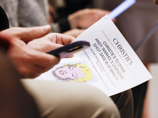 Eine linke Hand hält einen Flyer des Auktionshauses Christie's. Der Flyer zeigt das Bild "Shot Sage Blue Marilyn" von Andy Warhol. In der rechten Hand hält die Person ein Smartphone.