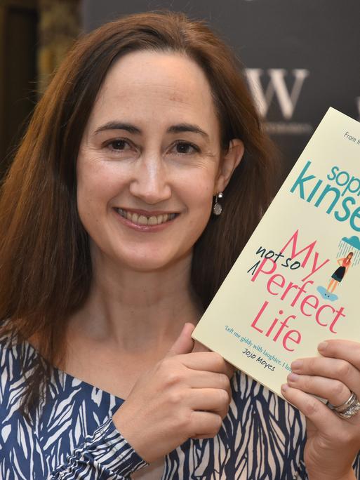 Die Autorin Sophie Kinsella hält ein Buch in die Hand und blickt in die Kamera.
