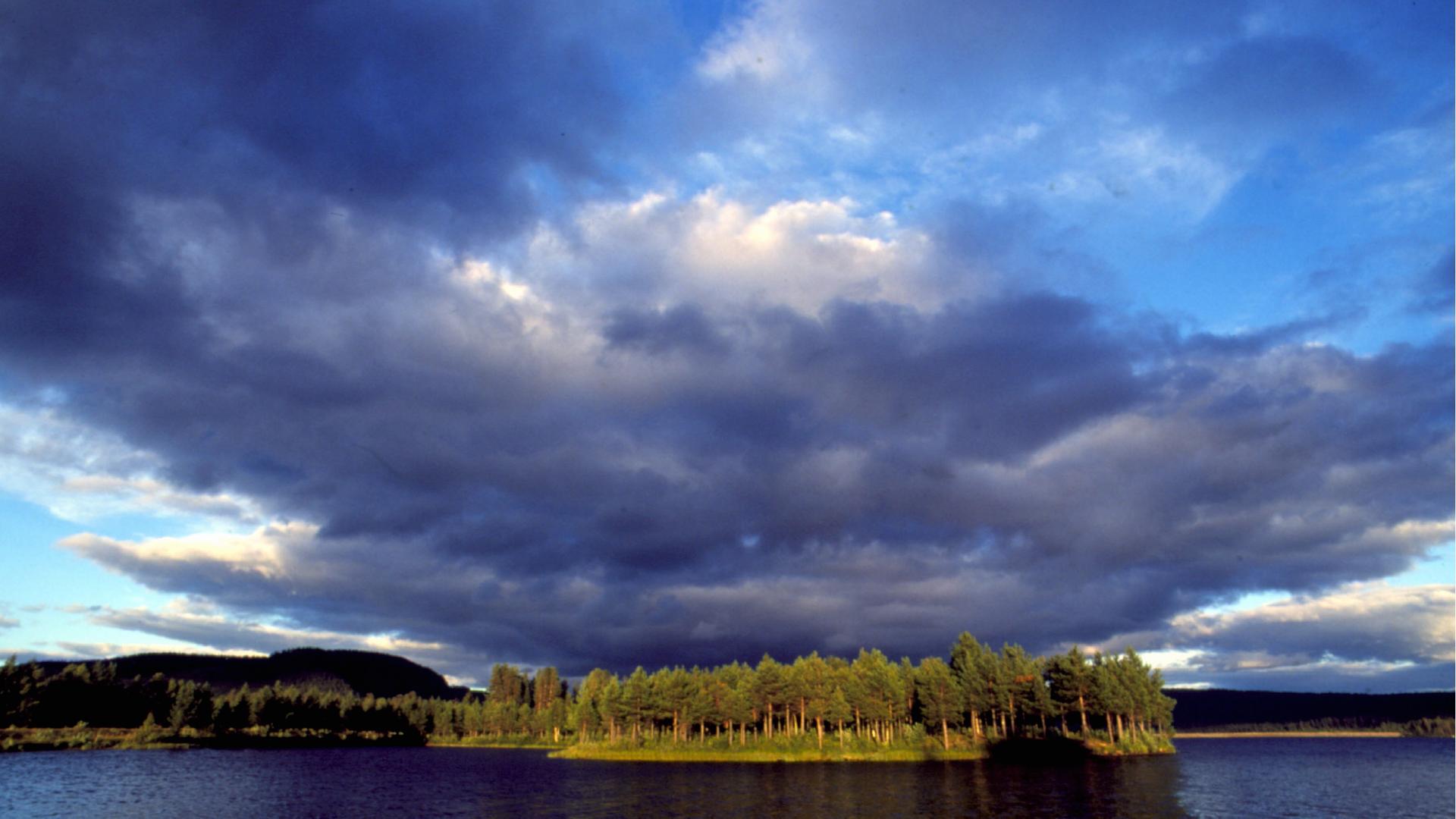 Über einem von Nadelbäumen gesäumten See türmen sich dunkle Wolken.