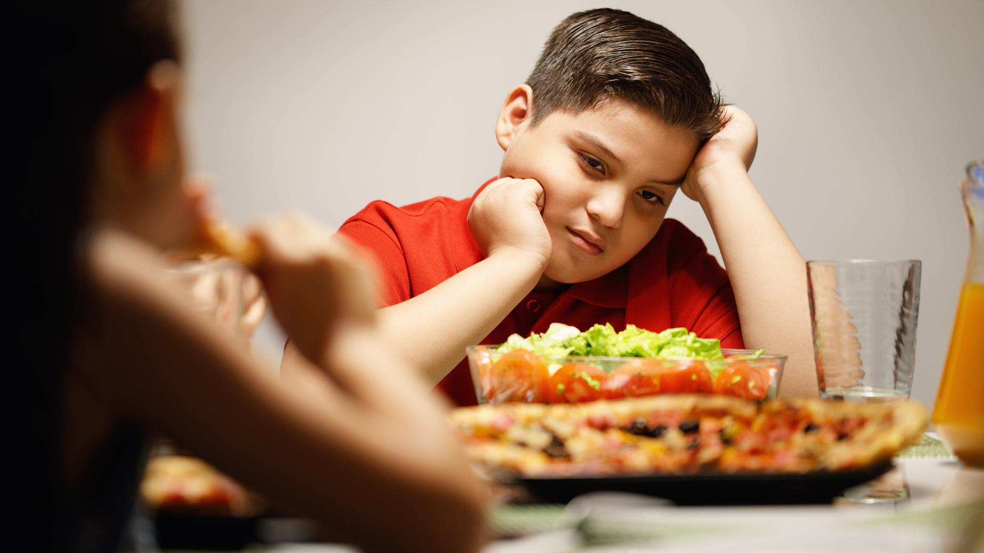 Ein junge sitzt am Essenstisch und stützt seinen Kopf auf seinen Arm. Im Vordergrund sind Pizza und Salat zu sehen.