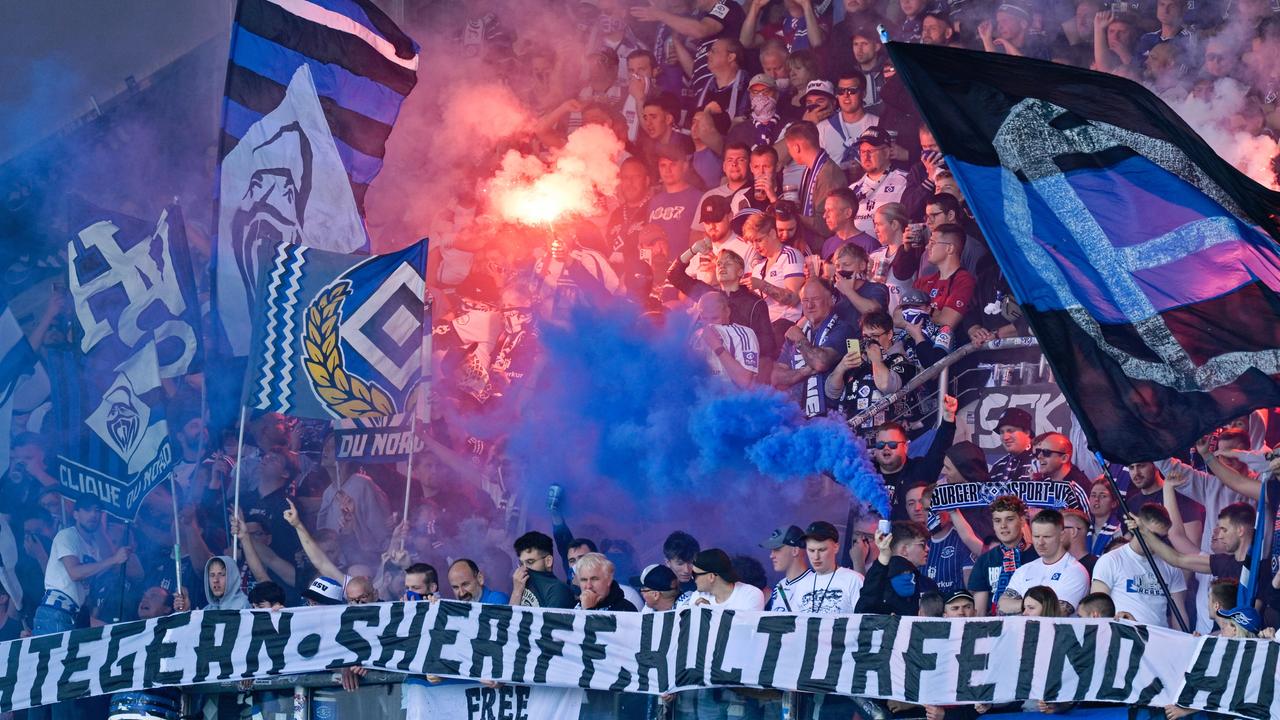 Hamburger Fans stehen mit Flaggen im Stadion. In der Menge wird Pyrotechnik gezündet.