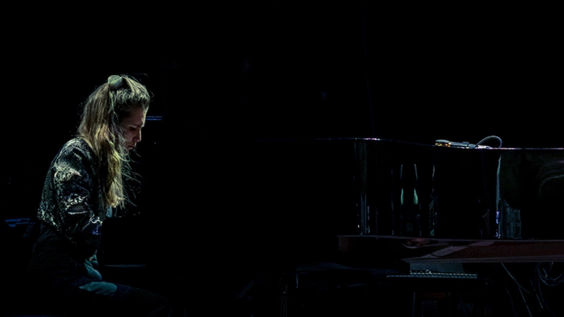 Eine junge Frau sitzt auf einer dunklen Bühne, nur sie und der Flügel sind schwach beleuchtet.