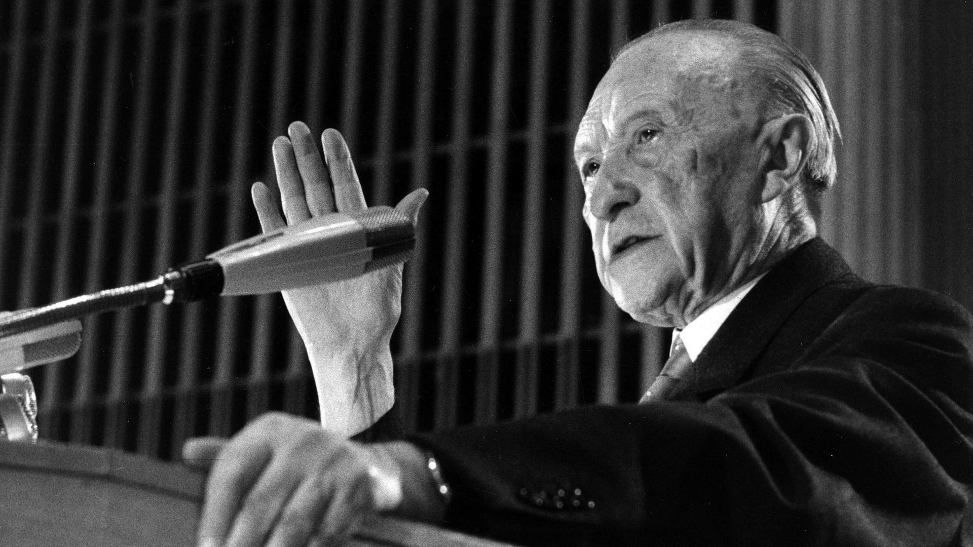Ein Schwarzweiß-Foto zeigt Konrad Adenauer von der Seite während er an einem Rednerpult in ein Mikrofon spricht und gestikuliert Ein Schwarzweiß-Foto zeigt Konrad Adenauer von der Seite während er an einem Rednerpult in ein Mikrofon spricht und gestikuliert