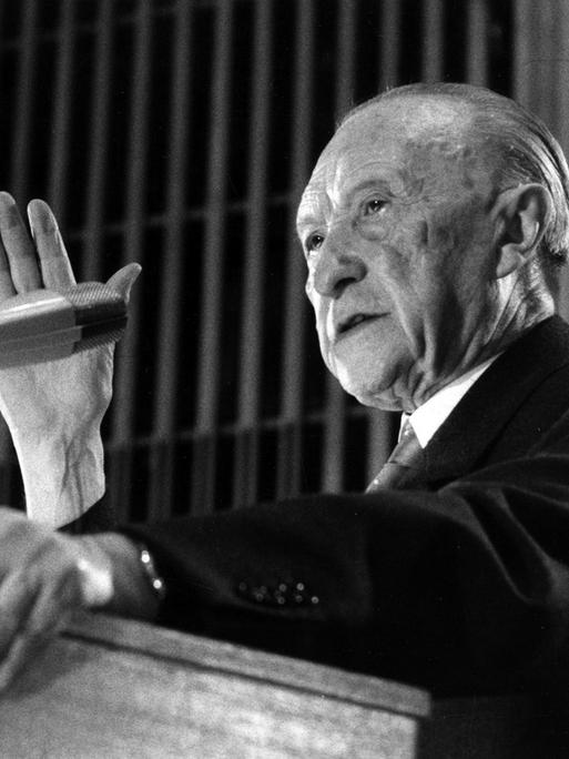 Ein Schwarz-Weiß-Foto zeigt Konrad Adenauer von der Seite, während er an einem Rednerpult in ein Mikrofon spricht und gestikuliert.